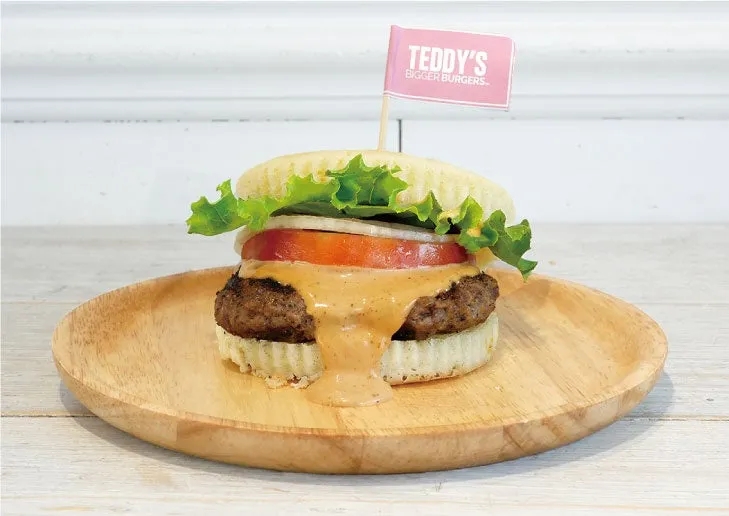 木製の皿に載ったハンバーガーの写真。 ハンバーガーは、レタス、トマト、玉ねぎ、ソース、パティ、バンズで構成されています。 ハンバーガーには、TEDDY