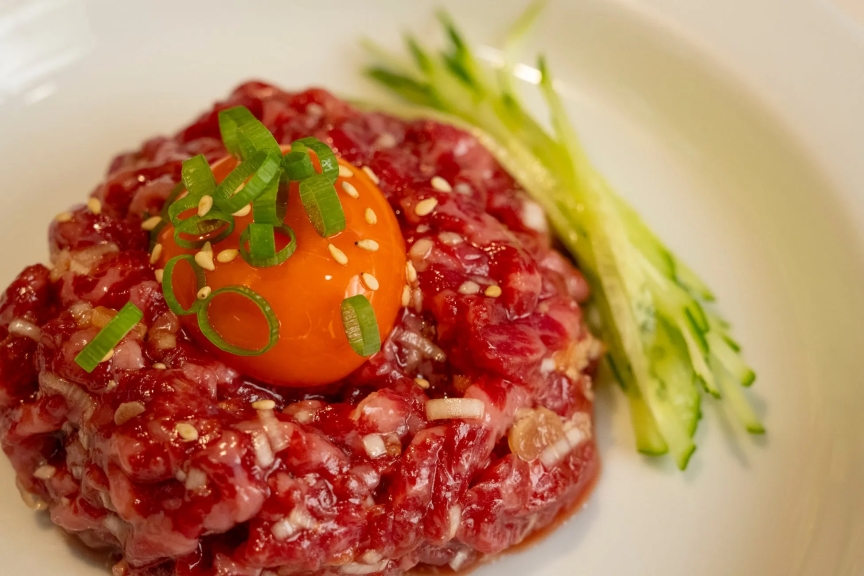 牛肉のタルタルに卵黄とネギがトッピングされ、キュウリが添えられた料理の写真です。