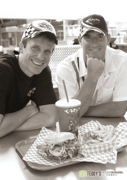 2人の男性がレストランでハンバーガーとフライドポテトを前に微笑んでいるモノクロ写真です。