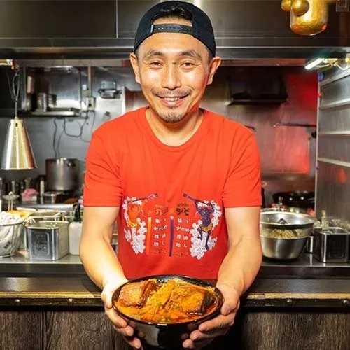 男性が料理を持っています。彼は赤いTシャツを着ていて、料理をカメラに向かって持っています。背景にはキッチンがあります。