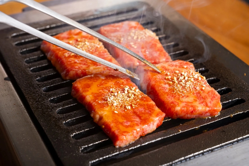 焼肉を焼いている様子を捉えた写真です。美味しそうな肉が鉄板の上で焼かれており、トングで掴んでいる様子も写っています。肉にはゴマがまぶされており、食欲をそそります。