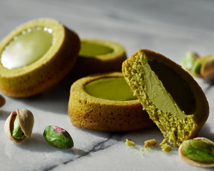 ピスタチオ風味の焼き菓子とピスタチオの実が置かれた写真です。焼き菓子は、外側が香ばしく、内側には鮮やかな緑色のクリームが詰められています。ピスタチオの実も添えられており、見た目にも美味しそうな印象を与えます。