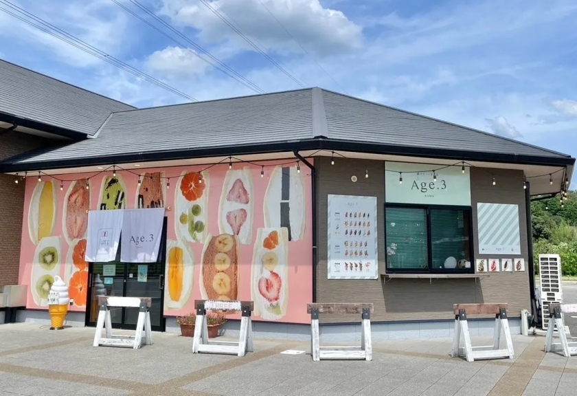 Age.3というお店の外観の写真です。お店の壁にはフルーツサンドのイラストが描かれており、メニューや看板も確認できます。お店の前にはテーブルとソフトクリームのオブジェがあります。