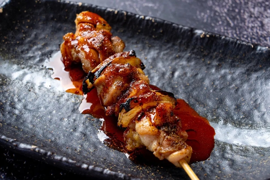 焼き鳥が一本、黒いお皿に盛り付けられています。串に刺さった鶏肉は照り焼きソースで覆われ、美味しそうに見えます。