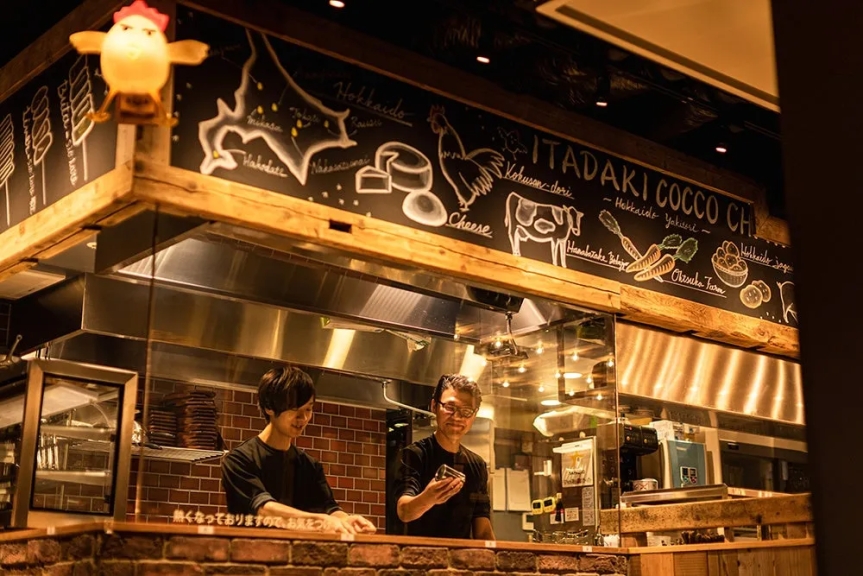 カウンター越しに料理をする2人の料理人と、黒板に書かれたメニューが印象的なお店の画像です。