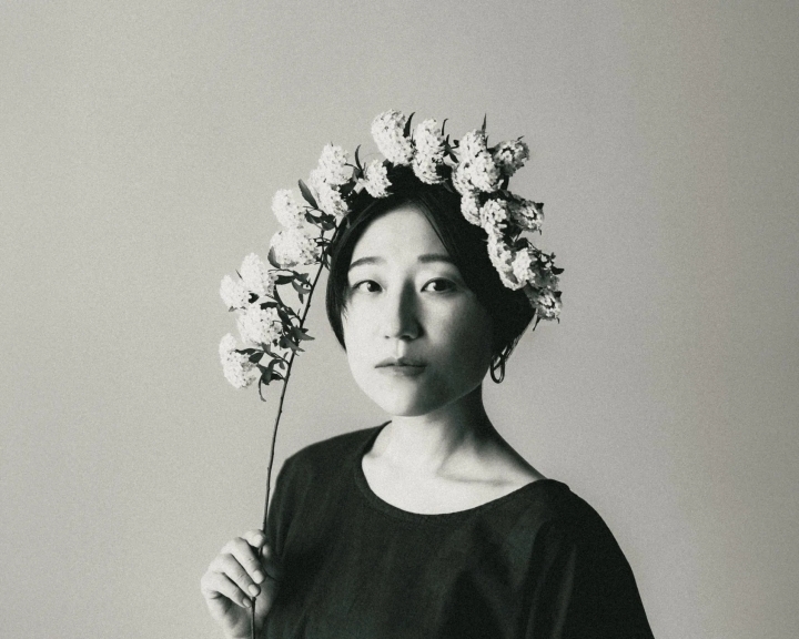 花冠をつけた女性のモノクロポートレート。女性は、白い花が咲いた枝を持ち、カメラ目線で少し悲しげな表情をしています。