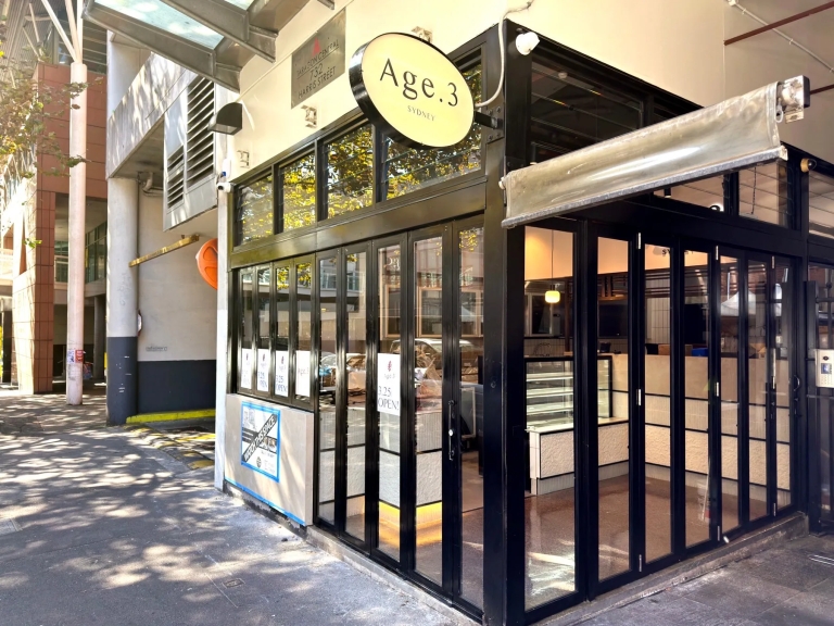 Age.3というカフェの外観写真。ガラス張りのドアと窓があり、店の看板には「Age.3 SYDNEY」と書かれています。店の前には歩道があり、周囲には建物が建っています。
