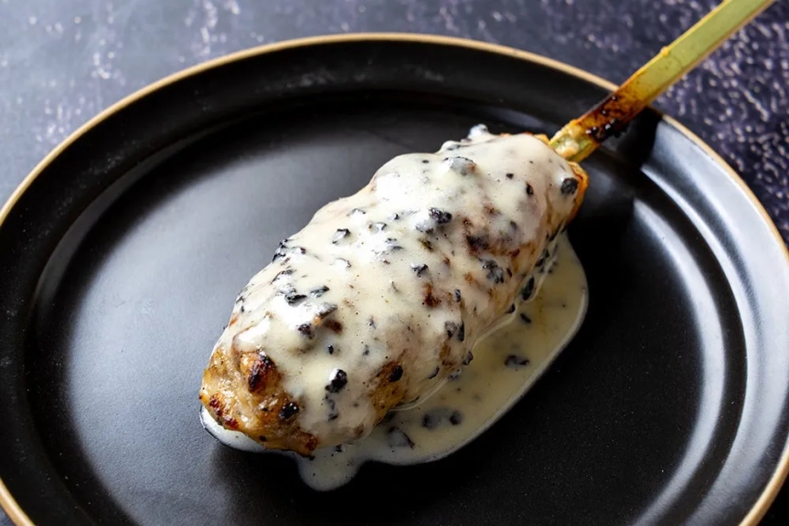 黒いお皿にのった、串に刺さった料理の写真です。料理は、白いソースで覆われています。ソースには黒い粒が見えます。料理は美味しそうです。