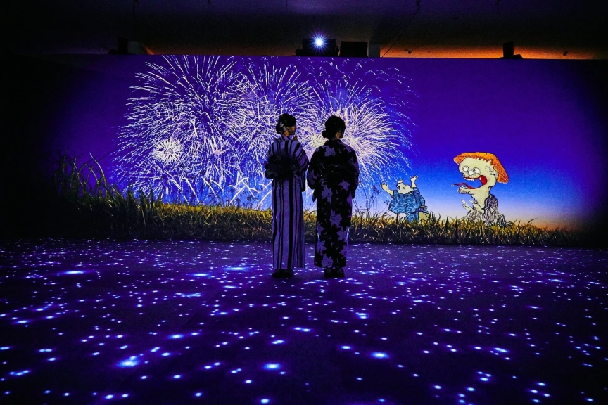 花火のプロジェクションマッピングを見ている二人の女性。足元には光の演出が施され、背景には妖怪のようなキャラクターも描かれている。