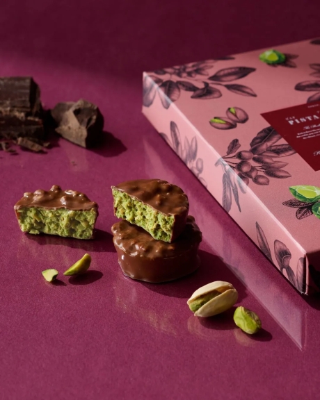 ピスタチオチョコレートのお菓子の写真です。チョコレートでコーティングされたピスタチオのお菓子がいくつか重ねられており、ピスタチオの実も添えられています。背景は紫で、黄色のエリアにブランド名が記載されています。