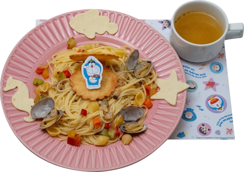 ピンクの皿に盛られたパスタとスープのセット。パスタには貝や野菜が添えられ、ドラえもんのキャラクターが飾られている。スープは白いカップに入っており、コースターにはドラえもんのイラストが描かれている。