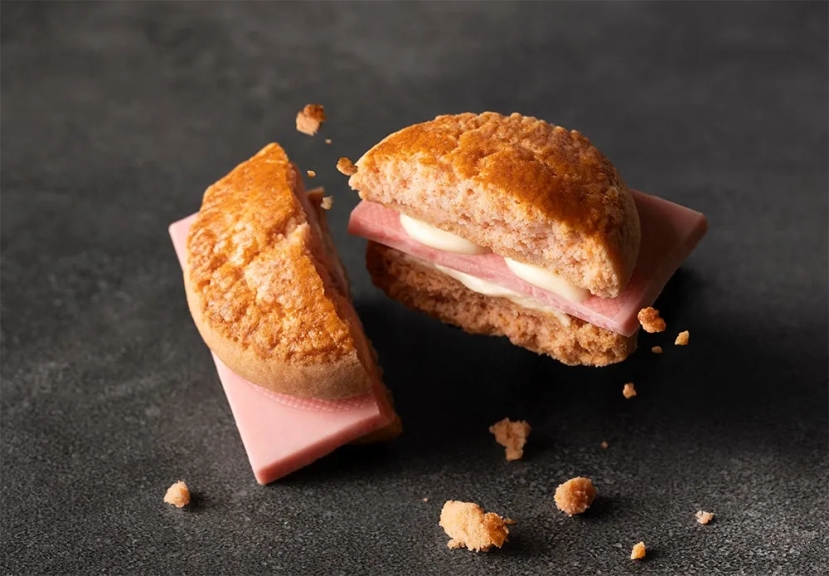 ピンク色のパンにハムと白いクリームが挟まれたサンドイッチの画像です。サンドイッチが半分にカットされており、パンの質感と中の具材がよく見えます。背景は暗い色調で、食べ物が際立っています。