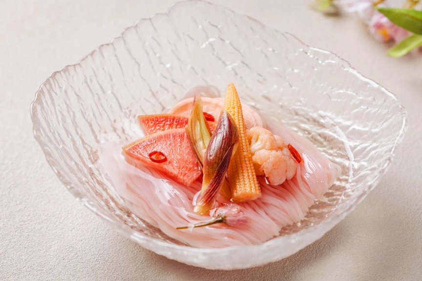 透明なガラスの器に盛られた、ピンク色の素麺と野菜の盛り合わせ。