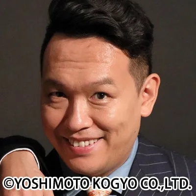 スーツを着たアジア系の男性がカメラに向かって笑顔を見せているクローズアップポートレートです。背景はシンプルで、顔と上半身が写っています。