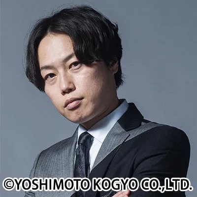 黒髪の男性がグレーと黒のツートーンスーツを着用し、カメラを真剣な表情で見つめているポートレート写真です。背景はシンプルなグレーです。