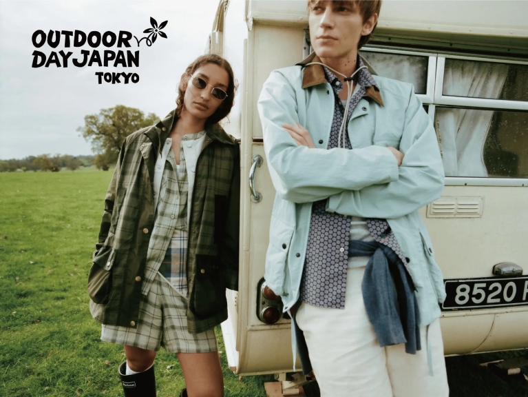 2人のモデルが屋外に立っており、一人は緑色のジャケットとサングラスをかけており、もう一人は水色のジャケットを着ています。背景にはキャンピングカーがあります。