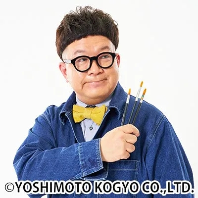 個性的な髪型と丸眼鏡をかけた男性が、黄色い蝶ネクタイとデニムジャケットを着用し、絵筆を手に微笑んでいます。アーティストのような雰囲気を持つ人物のポートレートです。