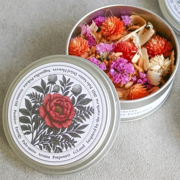 蓋が開いた金属製の容器に入ったポプリ。容器の蓋には、赤い花と葉のイラストが描かれており、周囲には文字が書かれています。容器の中には、オレンジ色と紫色のドライフラワー、木のチップ、その他のドライ素材が詰められています。